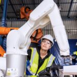 Genç kadın servis mühendisi, robot kontrol cihazını tutarak otomasyon fabrikasında robot kolu çalıştırıyor.